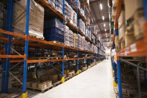 Interior de um armazém logístico amplo com corredores largos e prateleiras altas de estrutura metálica, carregadas com pallets contendo caixas e mercadorias embaladas.