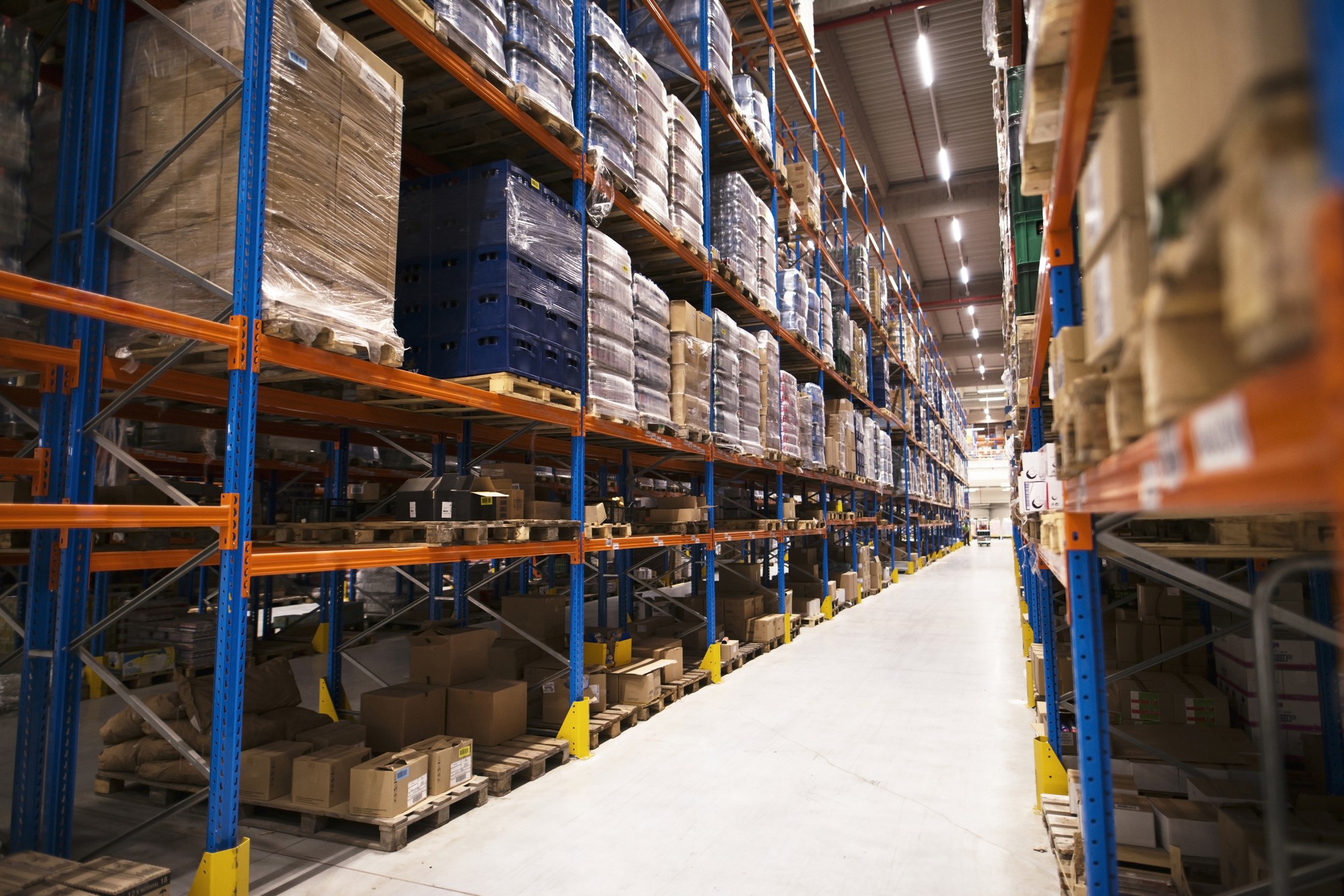 Interior de um armazém logístico amplo com corredores largos e prateleiras altas de estrutura metálica, carregadas com pallets contendo caixas e mercadorias embaladas.